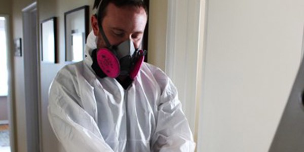 A person in a white protective suit and respirator mask adjusts their sleeve in the hallway during a mold investigation. The walls are adorned with framed pictures, and there's a door with a window in the background.