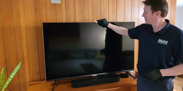 A person in a navy uniform and gloves is carefully sampling dust while cleaning a flat-screen TV on a wooden stand, surrounded by rich wooden paneling. Using a cloth, they inspect every corner of the screen, ensuring it's spotless as the TV remains turned off.