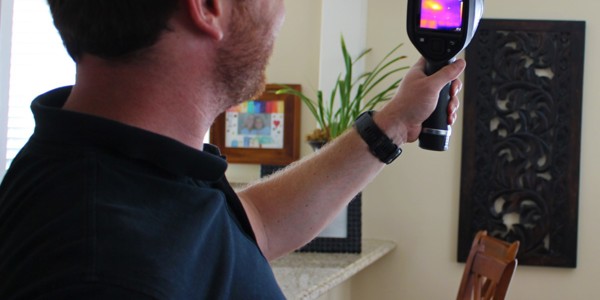 A man with glasses conducts an inspection using a handheld thermal imaging camera inside a room. The screen reveals a heat signature. He's wearing a dark shirt, and in the background, wooden chairs and a decorative wall hanging can be seen.